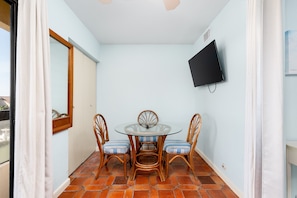 Uma pequena área de refeições com mesa de vidro e cadeiras de vime, televisão na parede e chão em mosaico.