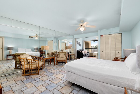 A hotel room with a large bed, wicker chairs, a ceiling fan, and a view of the ocean through the window.