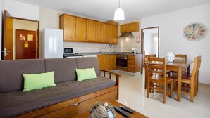 A living room with a sofa, wooden dining table, chairs, and a kitchen area with cabinets and appliances. - Apartment 'Campo Mar - T1 Beach View' with Sea View, Balcony and Wi-Fi (São Martinho do Porto)