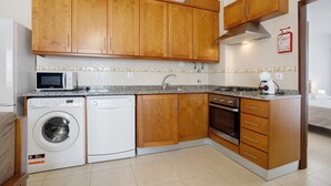 A compact kitchen with wooden cabinets, a microwave, a washing machine, and a stove. - Apartment 'Campo Mar - T1 Beach View' with Sea View, Balcony and Wi-Fi (São Martinho do Porto)