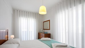 A hotel room with a bed, a nightstand, a dresser, a mirror, and sheer curtains. - Apartment 'Campo Mar - T1 Beach View' with Sea View, Balcony and Wi-Fi (São Martinho do Porto)