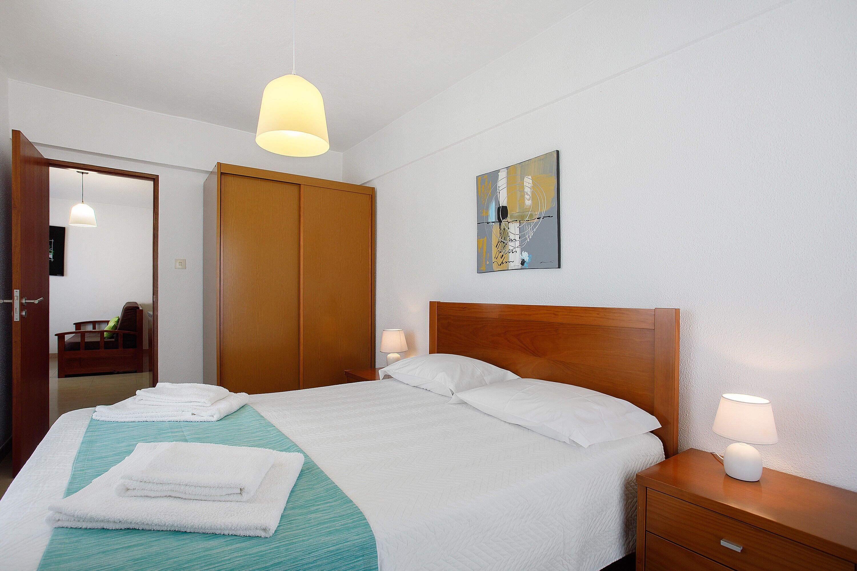 Un lit bien fait, avec du linge de lit et des serviettes blanches, une tête de lit en bois, une lampe de chevet et un tableau accroché au mur.