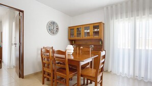A dining area with a wooden table and chairs, a clock, and a cabinet with glass doors. - Apartment 'Campo Mar - T1 Beach View' with Sea View, Balcony and Wi-Fi (São Martinho do Porto)