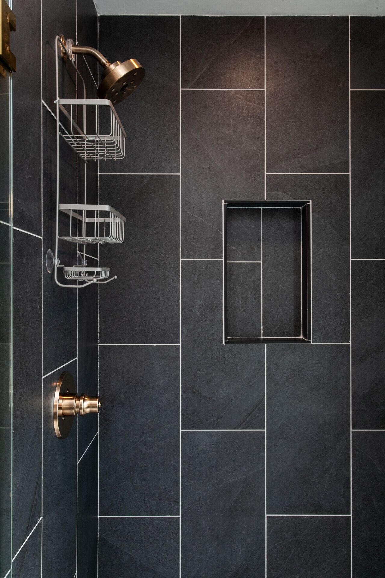 Ein modernes Badezimmer mit dunklen Fliesen, einer Dusche mit gebogener Brause und einer Metallregaleinheit.