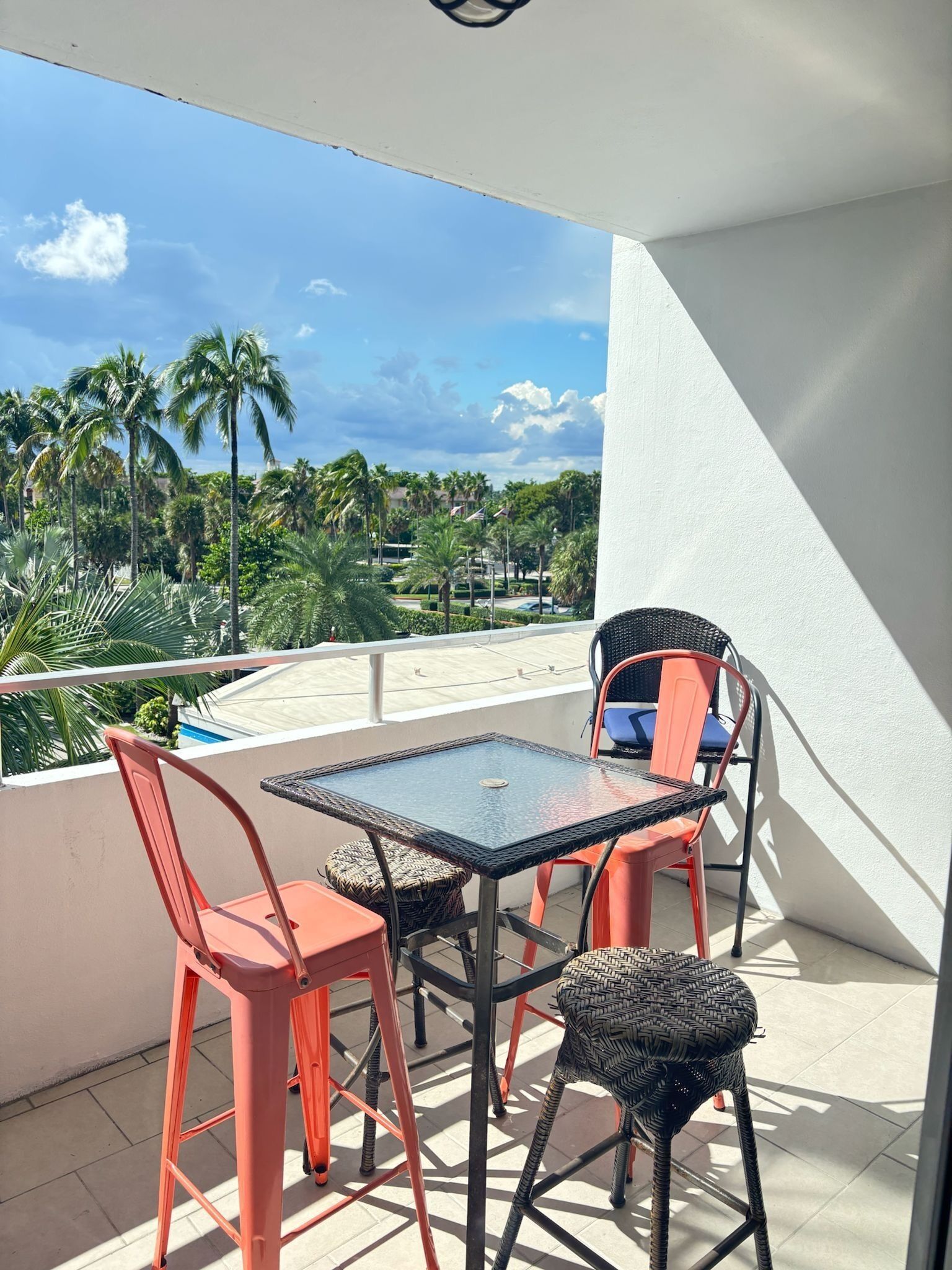 Un balcon avec une table et des chaises en verre, donnant sur une piscine et des palmiers.
