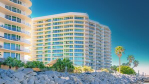A multi-story hotel with balconies, a rocky shoreline, and palm trees.