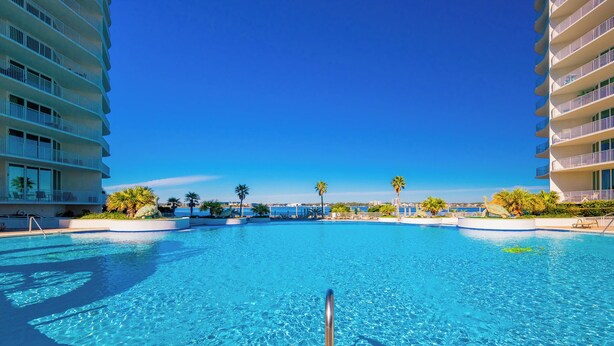 A large, clear swimming pool surrounded by palm trees and modern buildings.