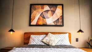 Un lit bien fait, avec une couverture matelassée et deux oreillers, une tête de lit en bois et, au-dessus, un tableau abstrait encadré.