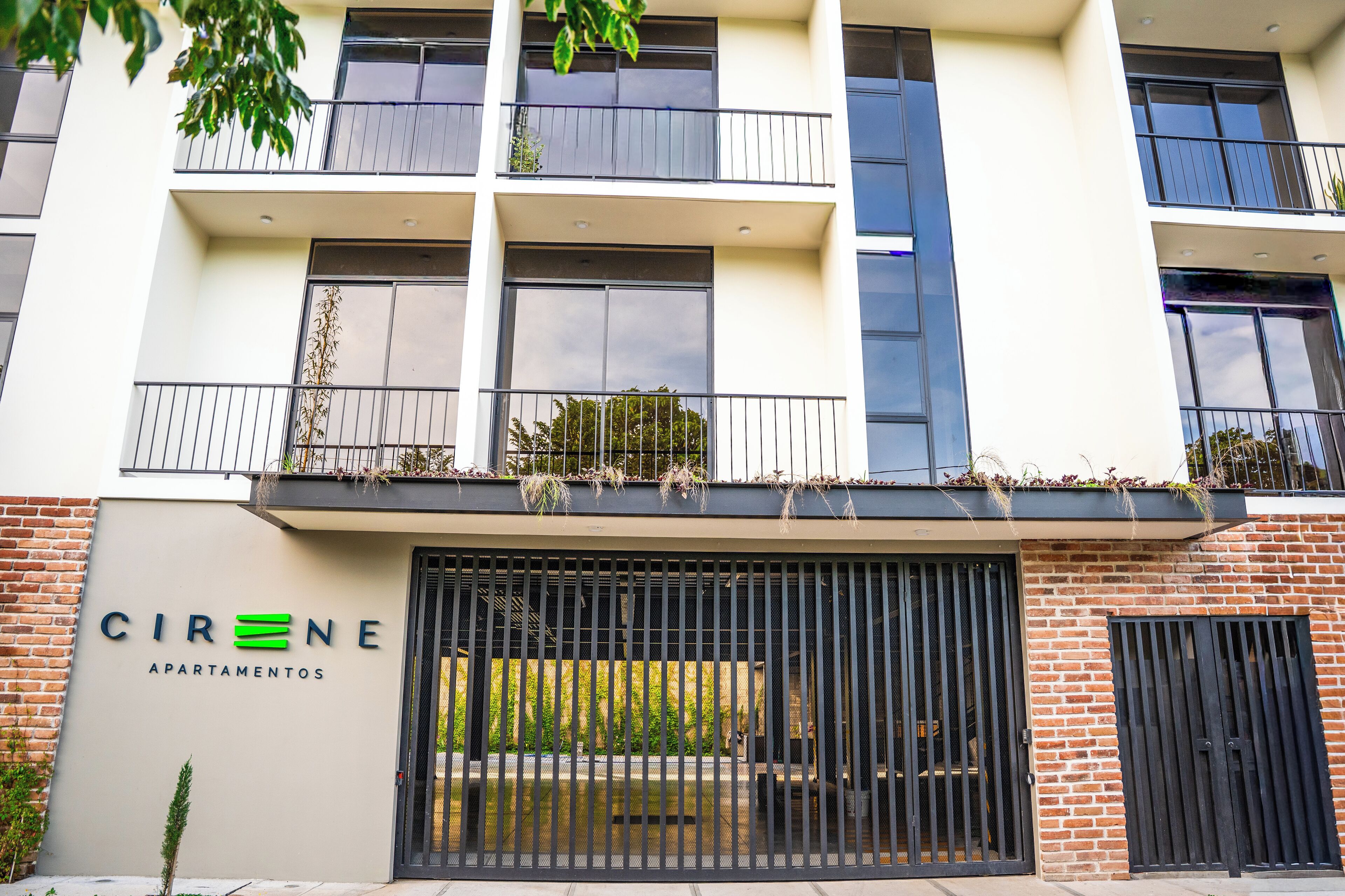 Un edificio de varios pisos con balcones, una entrada protegida por una reja y un cartel que dice 'CIRENE APARTAMENTOS'.