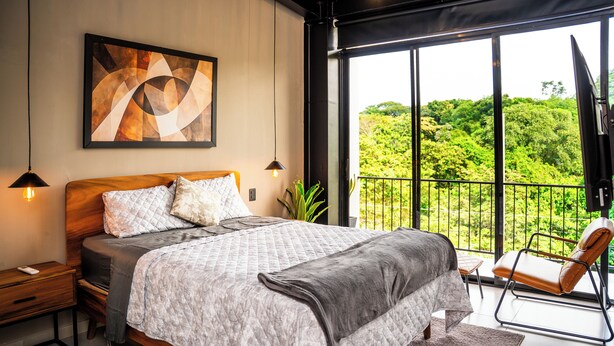 Une chambre dotée d’un grand lit, de tables de chevet, d’une commode, d’une chaise et d’un balcon offrant une vue sur la verdure.