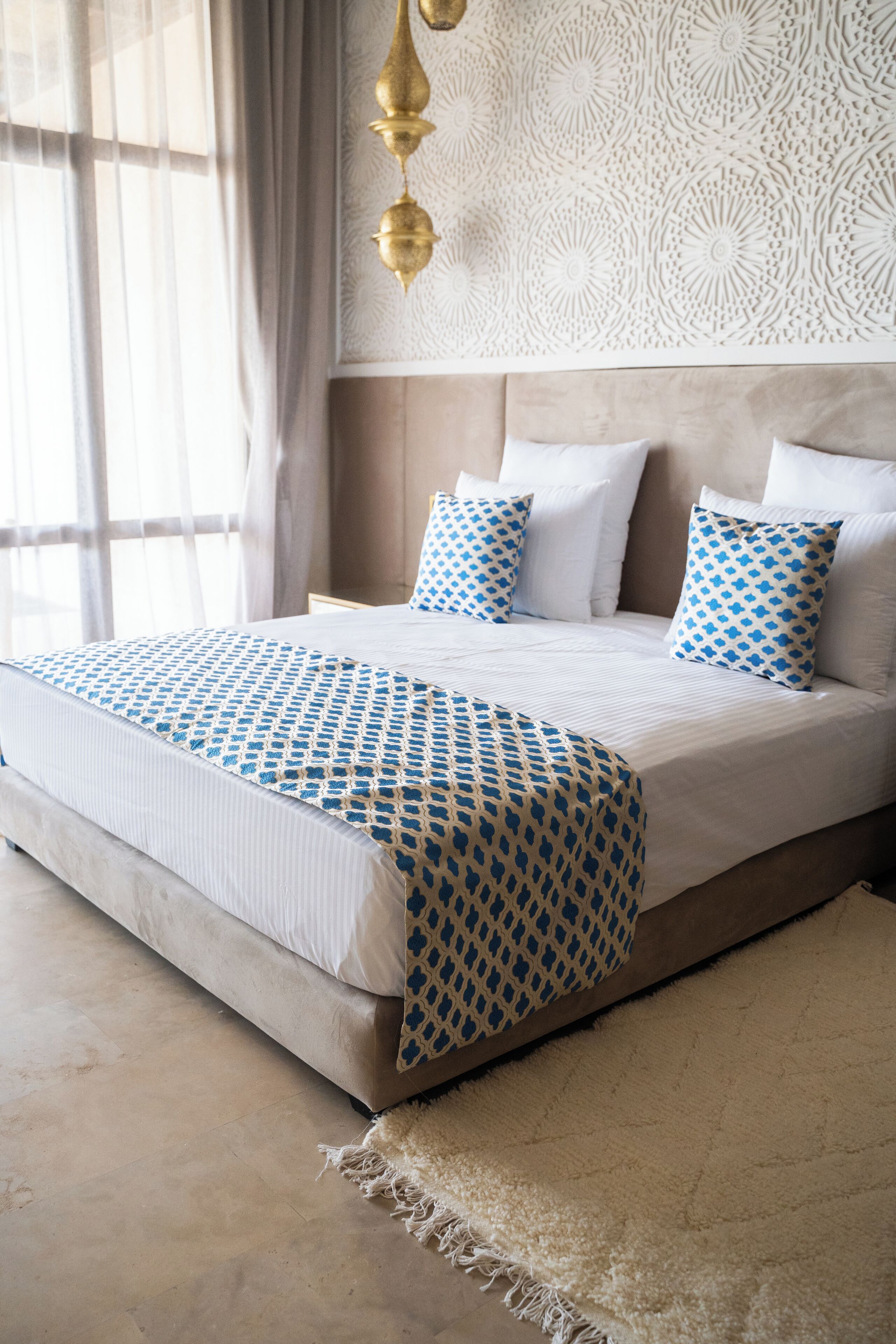 A bed with a patterned bedspread, a headboard with a decorative panel, and a light-colored rug.