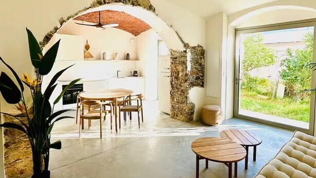 A spacious living area with a dining table and chairs, a sofa, a ceiling fan, and a large window offering a view of greenery. - Montera Apartment with Pool (Bastia)