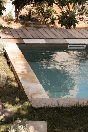 A rectangular swimming pool with a wooden deck and stone edging, surrounded by grass and trees.