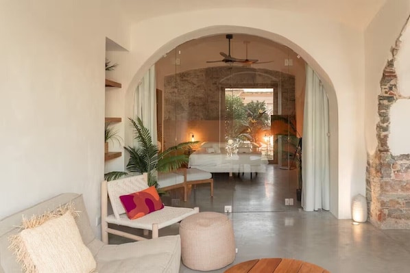 A modern living room with a sofa, armchair, and ottoman, a glass wall leading to a dining area, and a ceiling fan.
