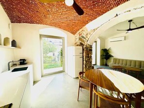 Cocina con encimera blanca, lavamanos y ventana. Zona de comedor con mesa y sillas de madera.