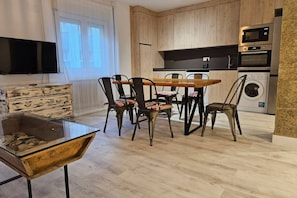 A modern kitchen with wooden cabinets, a dining table with chairs, a TV mounted on the wall, and a washing machine.