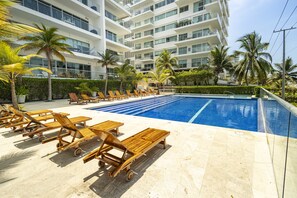 Uma área de piscina com espreguiçadeiras de madeira e palmeiras, rodeada por um edifício com várias andares e varandas.