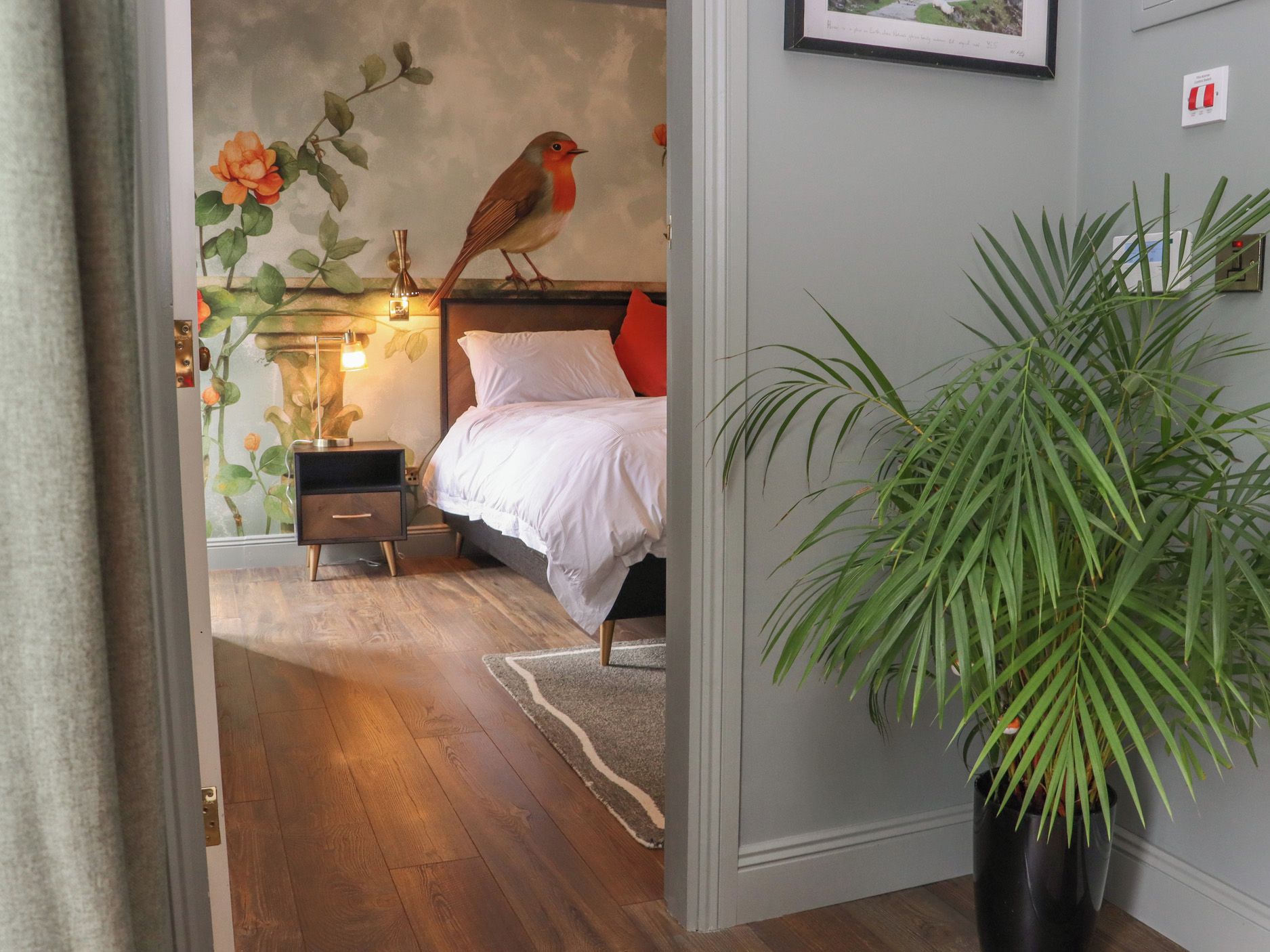 Quarto com cama, mesa de cabeceira, abajur e um quadro na parede de um pássaro.
