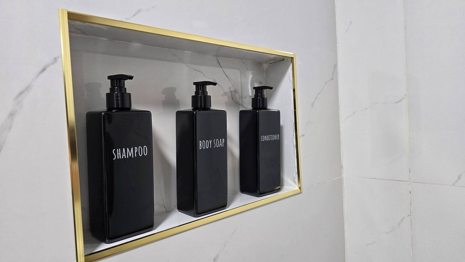 A bathroom with a marble wall and a shelf containing three black bottles labeled shampoo, body soap, and conditioner.