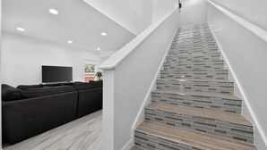 Un salon moderne avec un canapé d’angle noir, un téléviseur à écran plat et un escalier dont le revêtement présente un motif de carreaux original.