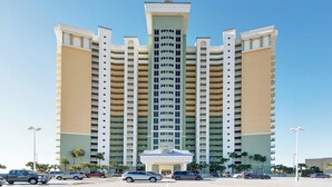 A multi-story hotel with a central tower, multiple balconies, and a parking lot with cars.