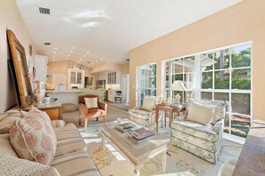 A spacious living room with a large window, a kitchen in the background, and a cozy seating area with patterned cushions.
