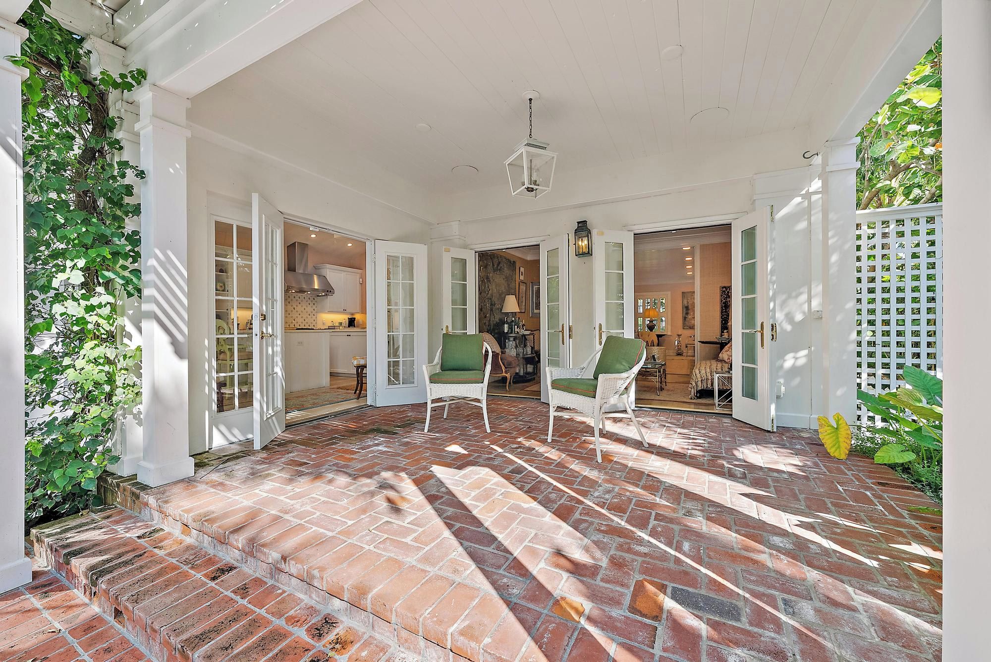 A covered patio with brick flooring, white columns, and two wicker chairs with green cushions.