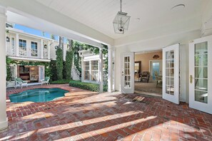A spacious indoor area with a brick floor, a pool, and a view of a house with a balcony.