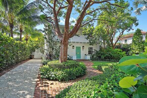 A house with a brick pathway leading to the front door, surrounded by lush greenery and palm trees.