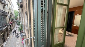 A narrow street with buildings on either side, a balcony with hanging plants, and a glimpse of a hotel room with a bed and a bathroom visible through a window.
