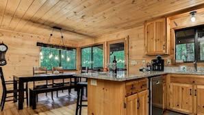 Interior de una cabaña de madera con una cocina que incluye una isla de madera, una mesa de comedor con bancos y grandes ventanales con vistas a la vegetación.