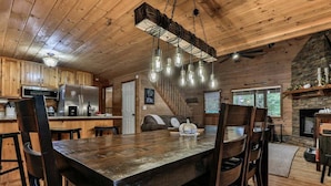 Interior rústico de una cabaña con mesa y sillas de madera, zona de cocina, chimenea y escalera.