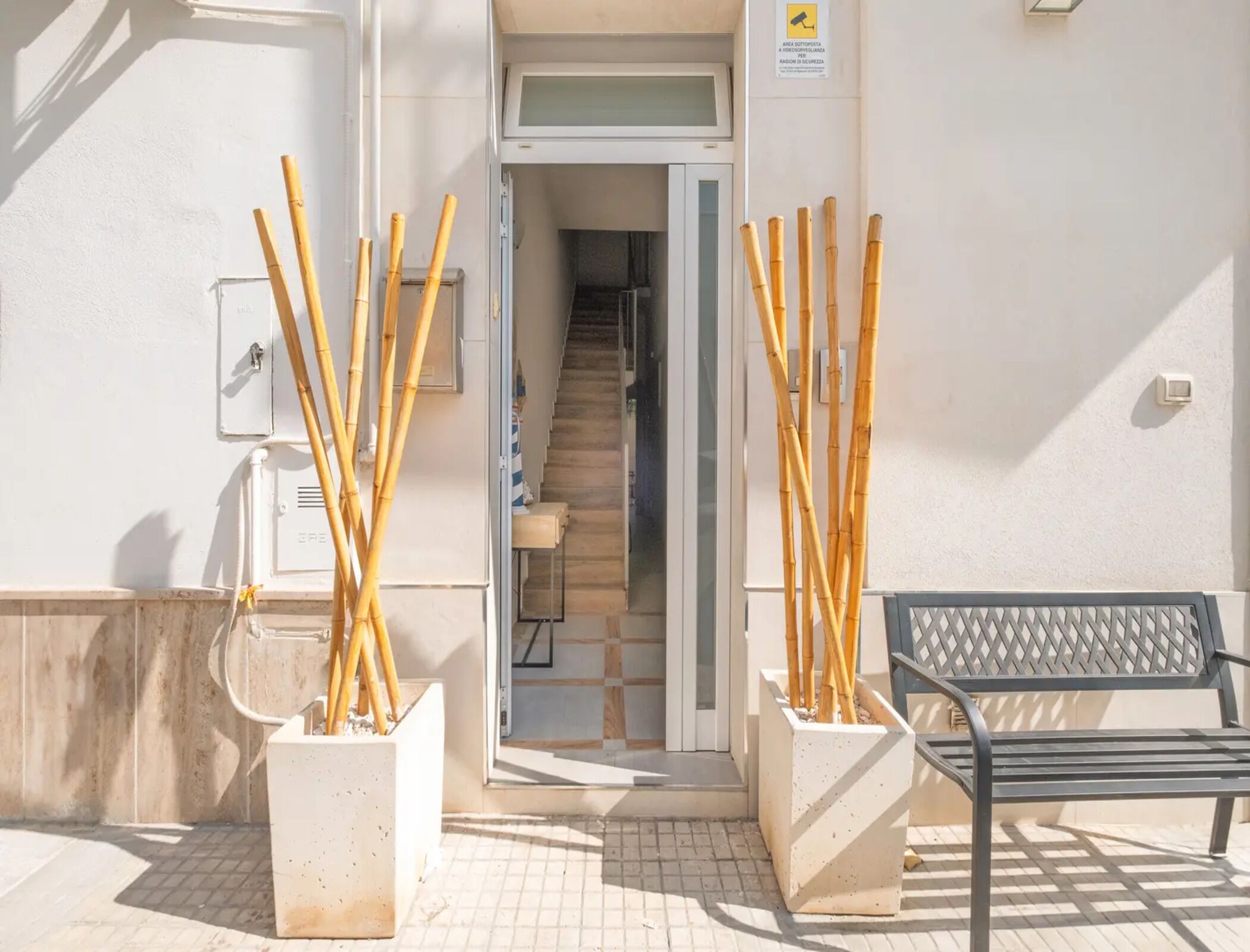 A building entrance with a staircase, two planters with bamboo sticks, a bench, and a door with a sign.