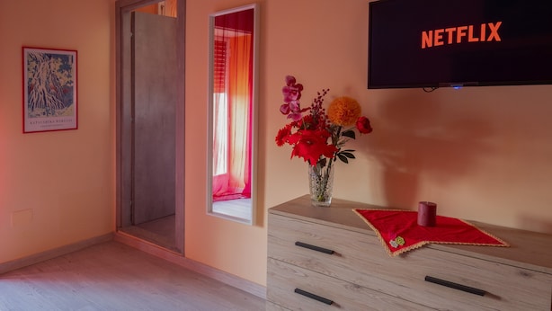 A room with a wooden dresser, a vase with flowers, a framed artwork, a sliding door, and a TV displaying 'NETFLIX'.