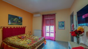 A bedroom with a bed, a window with curtains, a radiator, a wall-mounted TV, and a vase with flowers.