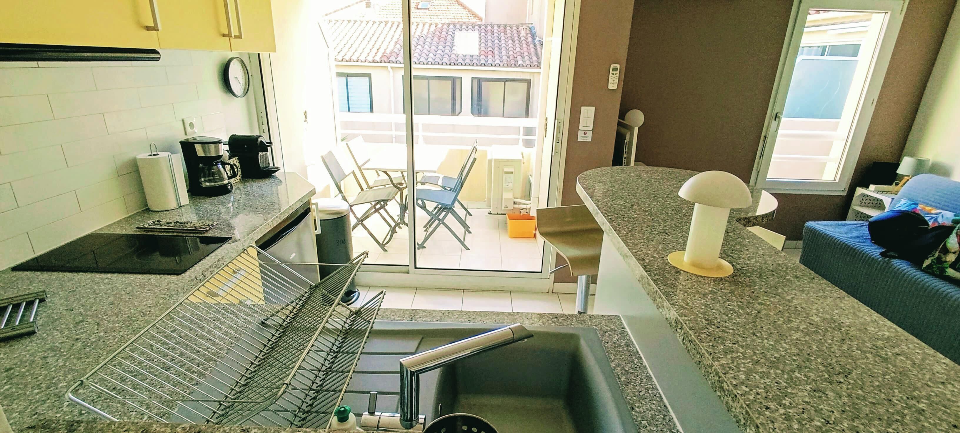 Eine Küche mit Spüle, Arbeitsfläche und Stühlen. Ein Fenster bietet Blick auf Gebäude.