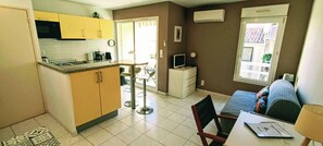 A compact kitchen with yellow cabinets, a microwave, and a coffee maker. There is a dining area with a table and chairs, and a TV on a small stand.