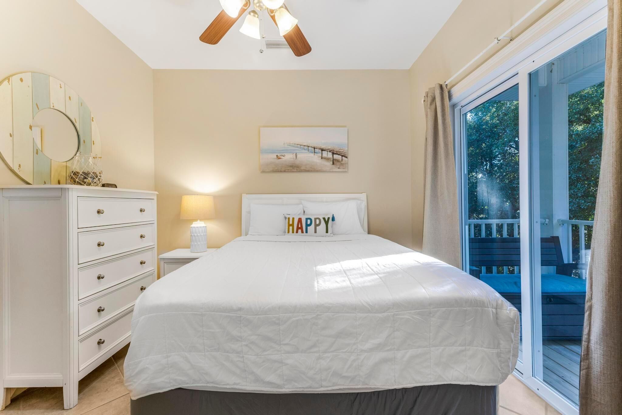 Un dormitorio con cama, cómoda, mesita de noche, lámpara, ventilador de techo, un cuadro y vistas a una terraza a través de una ventana.
