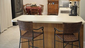 A kitchen with a central island, two wicker chairs, a wooden cabinet, a refrigerator, and a ceiling fan.