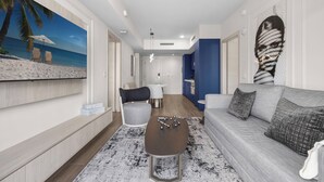 Un salon moderne avec une fresque murale représentant une plage, un canapé gris, une table basse et un coin repas avec une table et des chaises blanches.