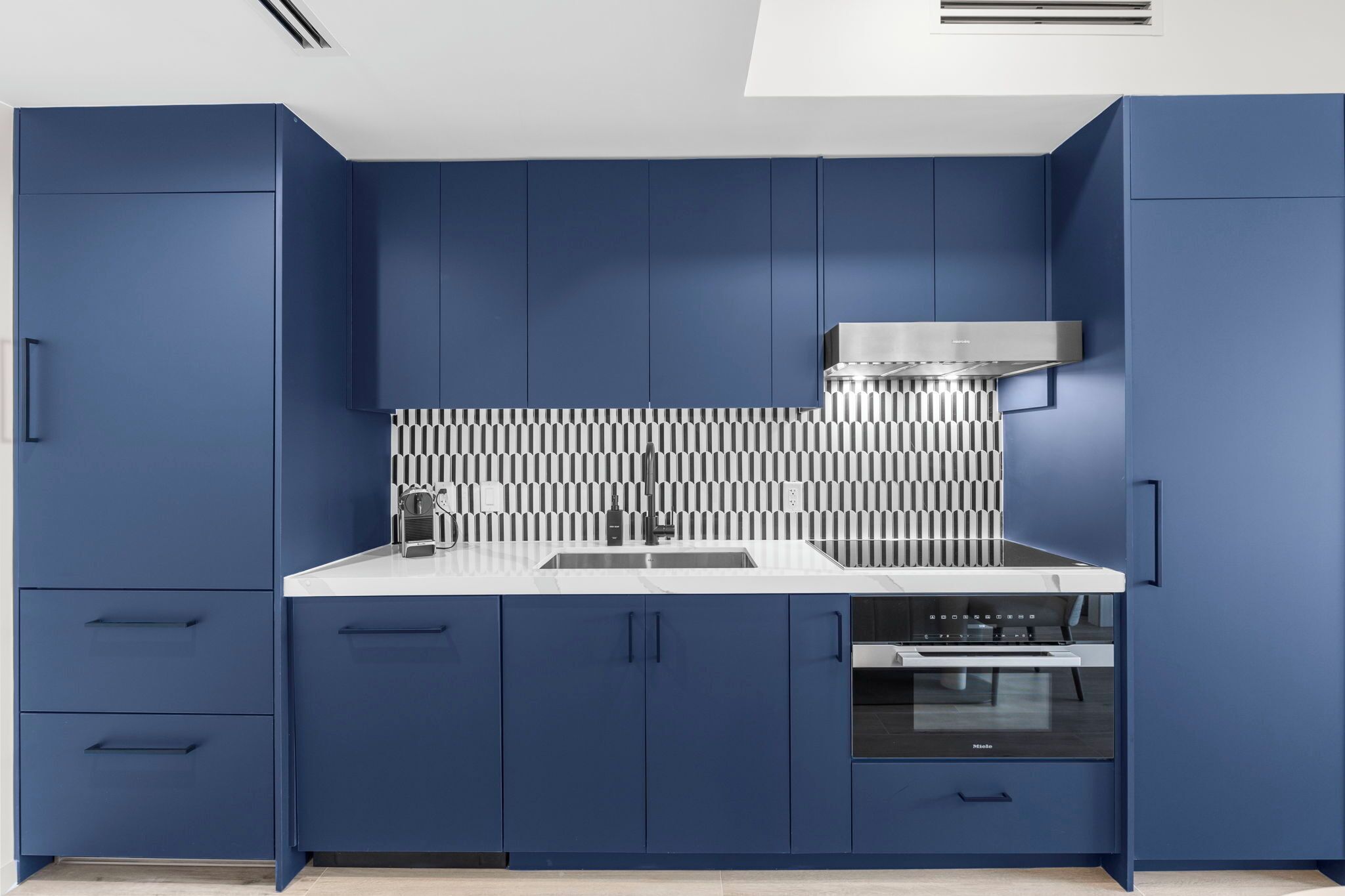 A modern kitchen with blue cabinets, a white countertop, a stainless steel range hood, and a built-in oven.