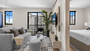 Un salon moderne avec un canapé gris, une table basse et un lit. Une grande fenêtre offre une vue sur les bâtiments et donne accès à un balcon.