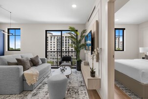 Una sala de estar moderna con un sofá gris, una mesa de centro y una cama. Hay un ventanal con vistas a edificios y un balcón.