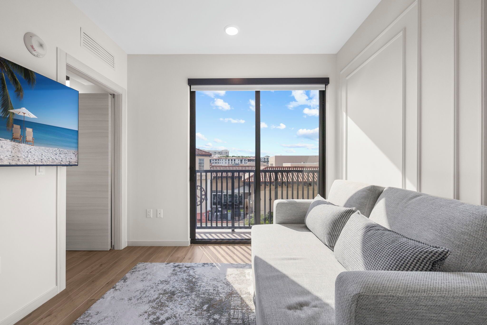 A modern living room with a grey sofa, a painting of a beach scene, and a large sliding glass door leading to a balcony with a city view.