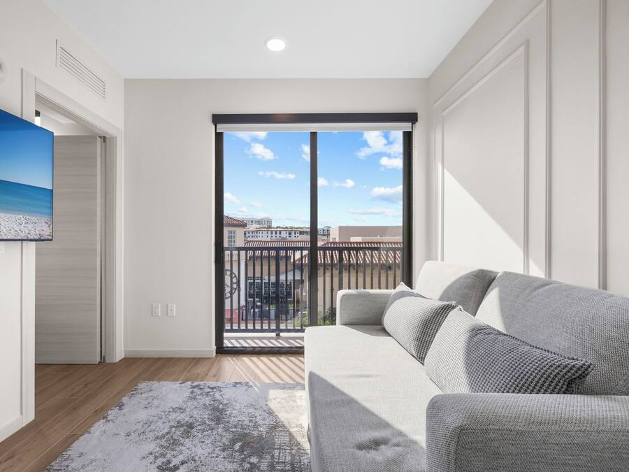 A modern living room with a grey sofa, a painting of a beach scene, and a large sliding glass door leading to a balcony with a city view.