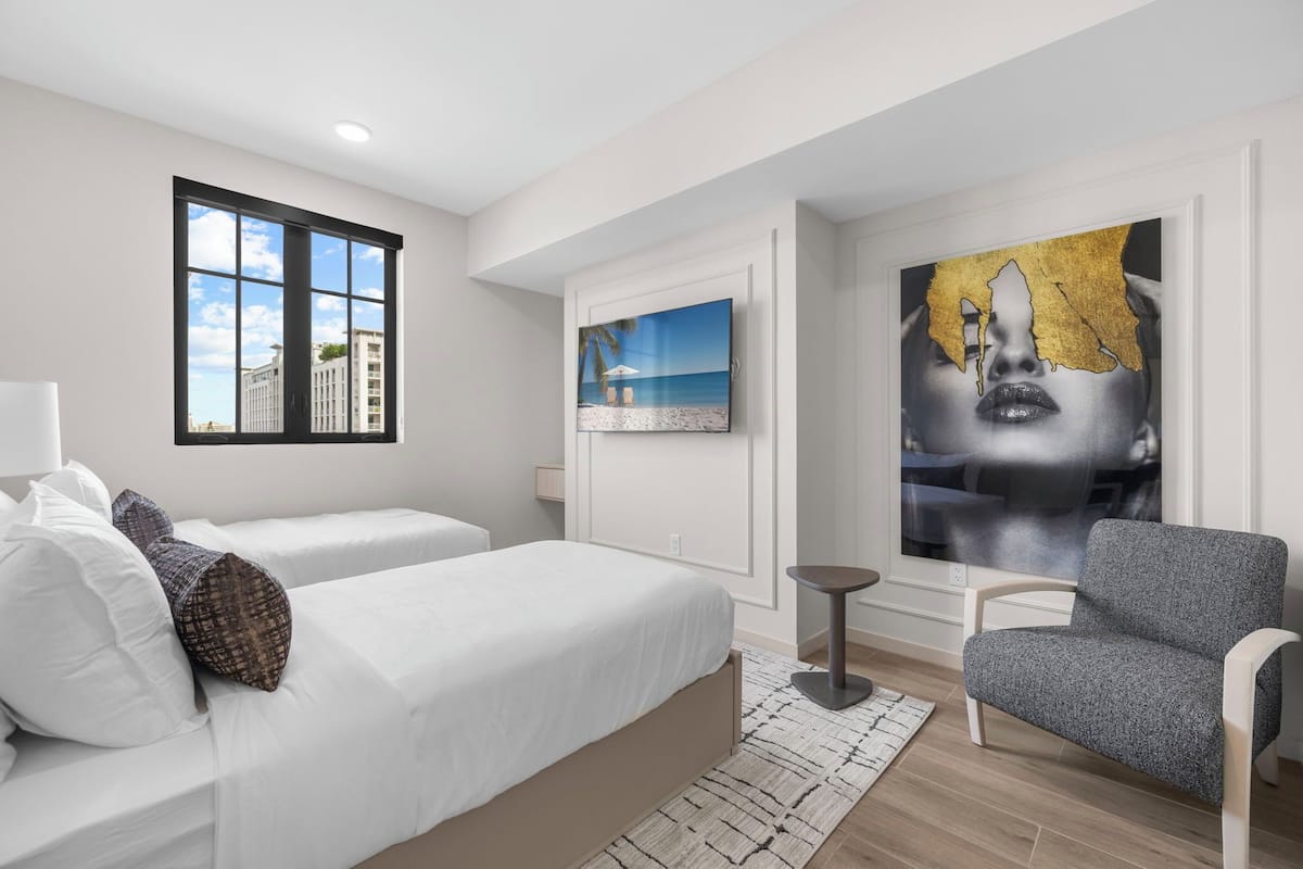 Ein modernes Hotelzimmer mit großem Fenster, einem Bett mit weißer Bettwäsche, einem grauen Sessel und einem an der Wand montierten Fernseher.