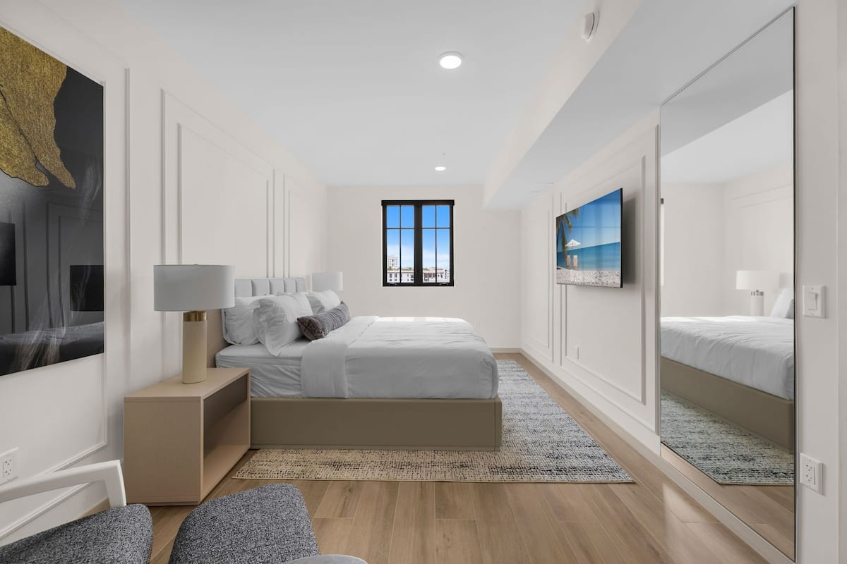 Ein modernes Schlafzimmer mit einem großen Bett, einem Nachttisch, einem Spiegel und Blick auf die Stadt durch ein Fenster.