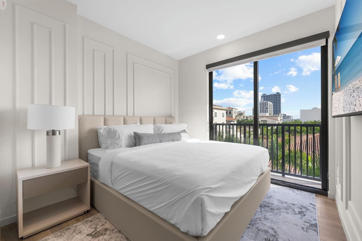 Ein Schlafzimmer mit einem großen Bett, einem Nachttisch, einer Lampe und Blick auf die Stadt vom Balkon aus.