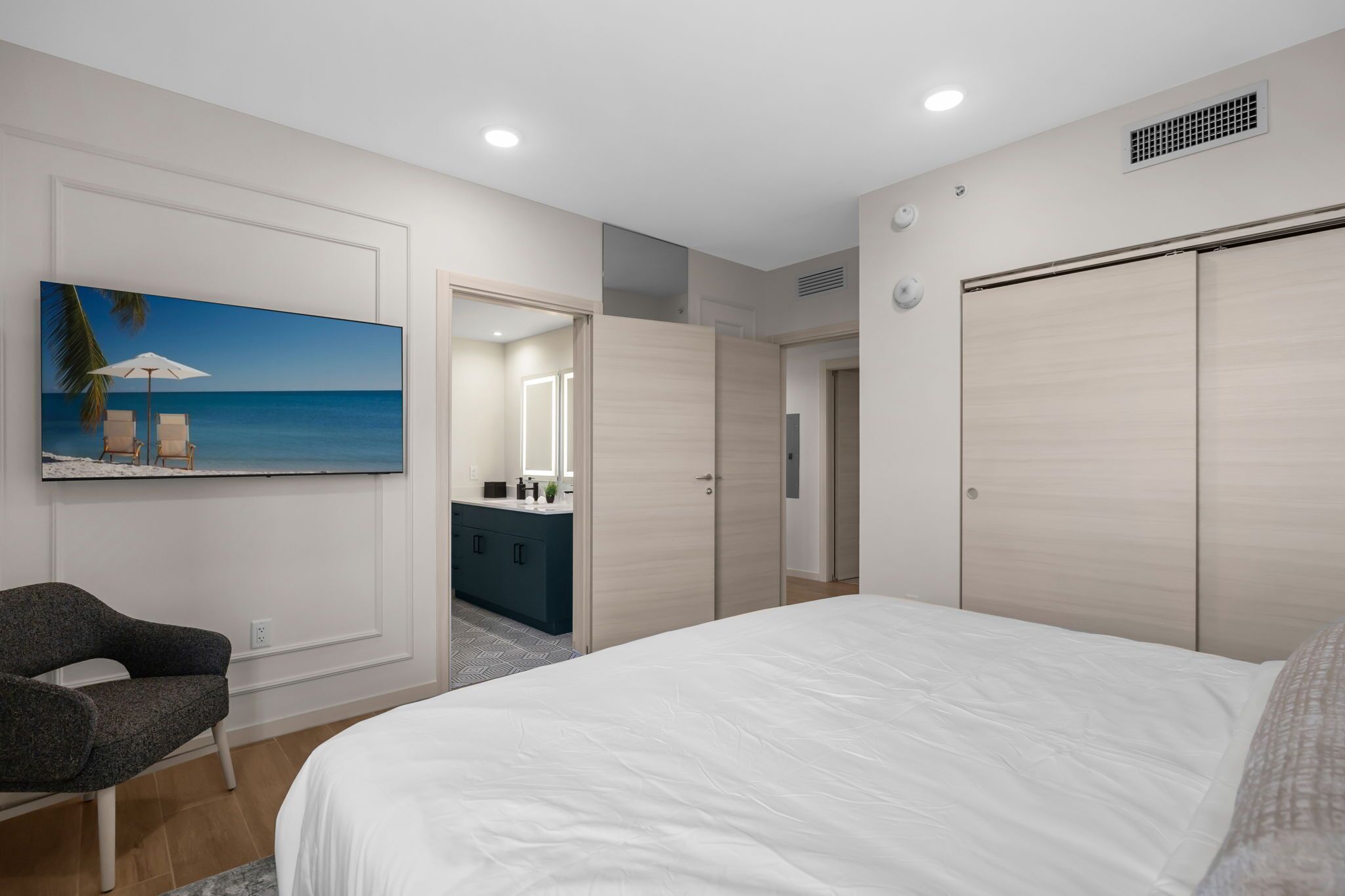 Une chambre à coucher avec un grand lit, une chaise, un tableau représentant une plage, une coiffeuse avec un miroir et une armoire.