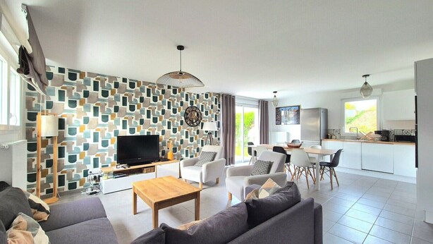 Un salon moderne avec un canapé, une table basse et un téléviseur. On y trouve également un coin repas avec des chaises et une table. La cuisine est visible, avec un réfrigérateur et une fenêtre.